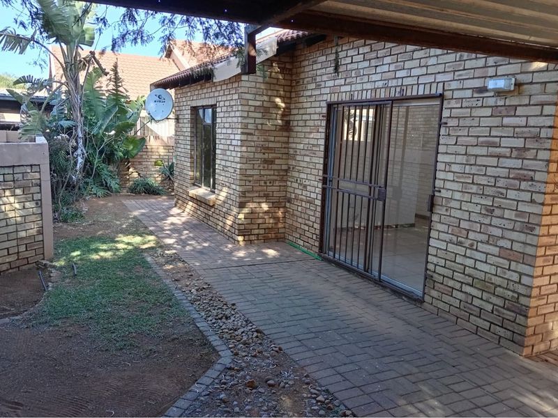 Rear exterior view of a face brick townhouse featuring a paved walkway, covered patio area, and sliding glass door with security gate.