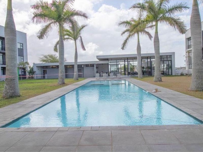 A well-maintained rectangular swimming pool set within a landscaped garden area, featuring stone coping and a modern clubhouse building in the background.