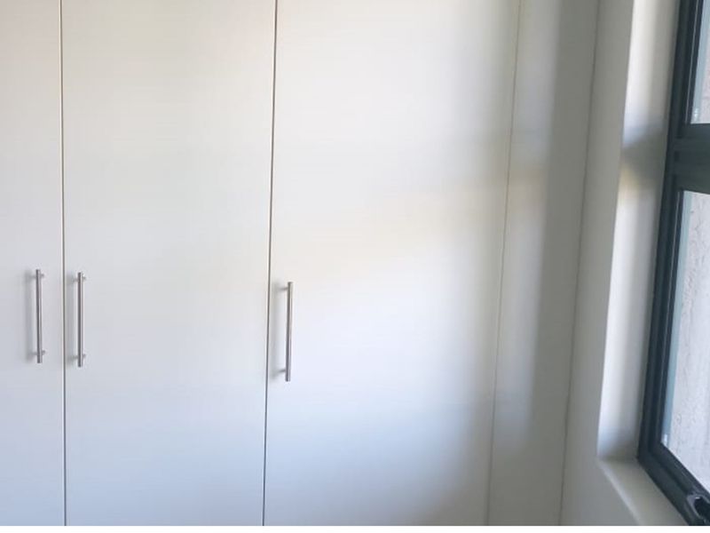 A clean, minimalist bedroom space featuring white built-in cupboards with modern metal handles and a side window.