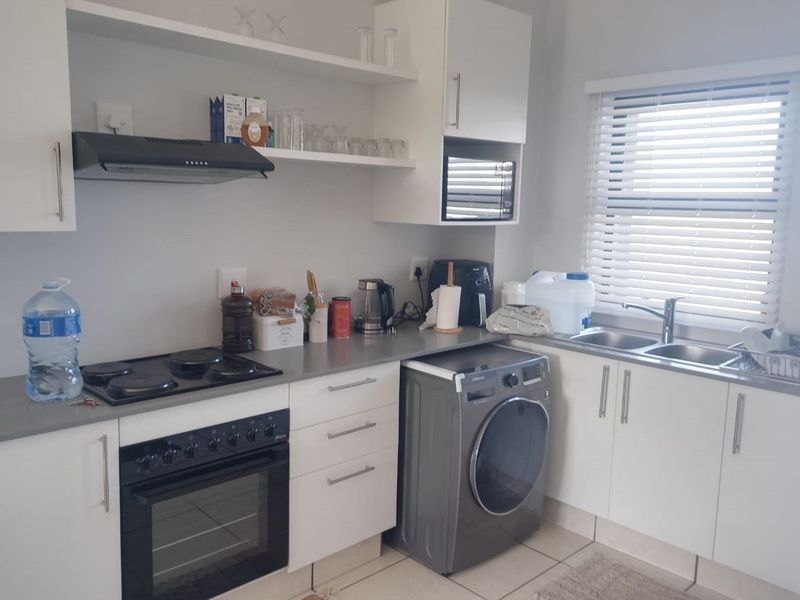 A functional kitchen featuring white cabinetry, grey countertops, an electric hob, built-in oven, extractor fan, and a double sink with venetian blinds.