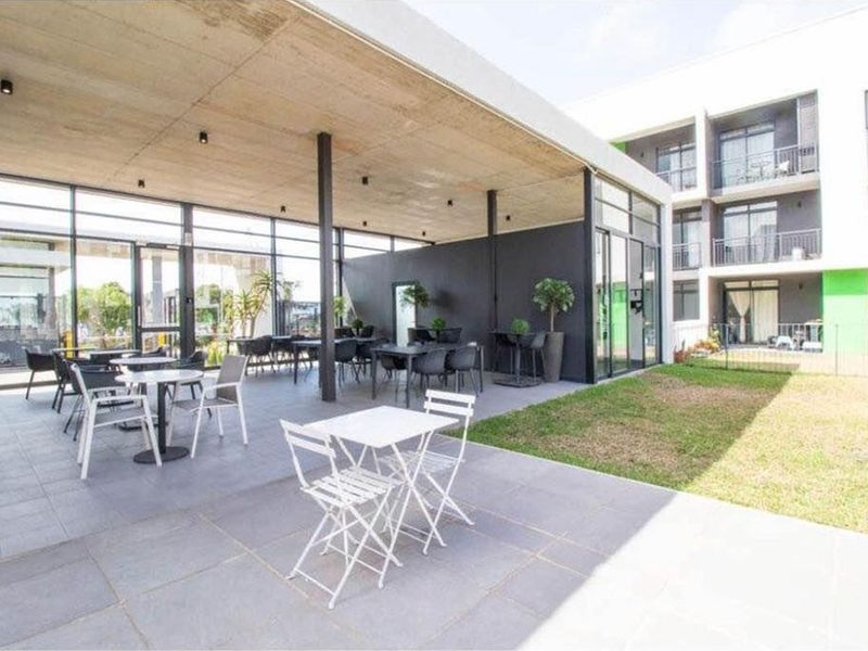 A modern covered patio area featuring a concrete ceiling, floor-to-ceiling glass walls, and tiled flooring, overlooking a communal garden space.