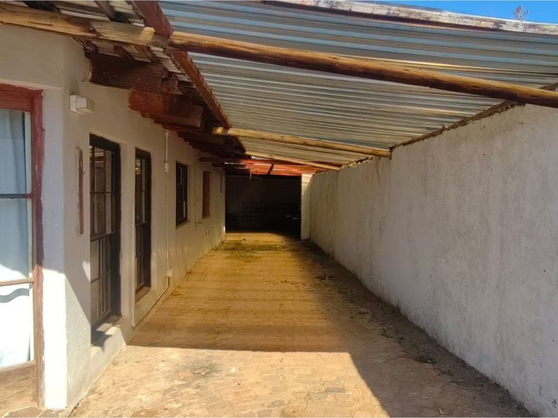 A narrow, covered outdoor walkway or side passage featuring a corrugated iron roof supported by timber beams and paved flooring.