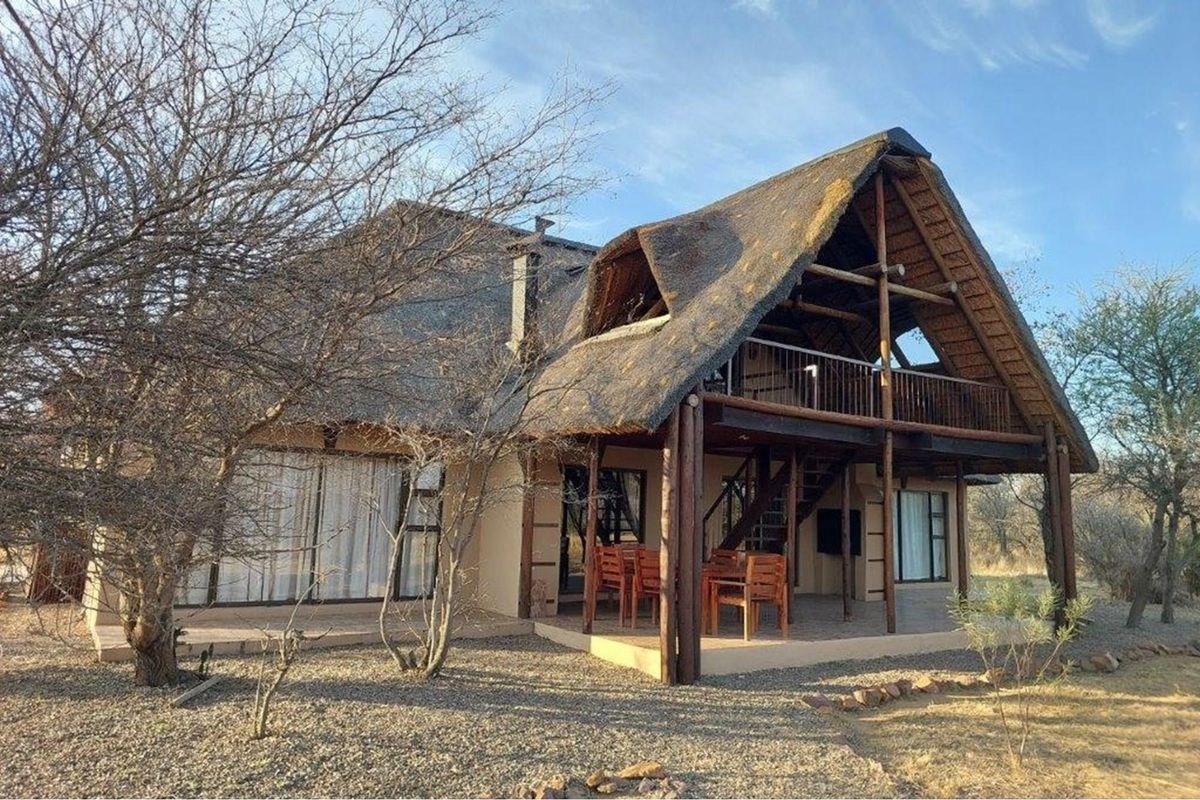 A charming double-story thatched-roof house featuring a spacious covered patio, wooden balcony, and rustic architectural design set in a natural landscape.