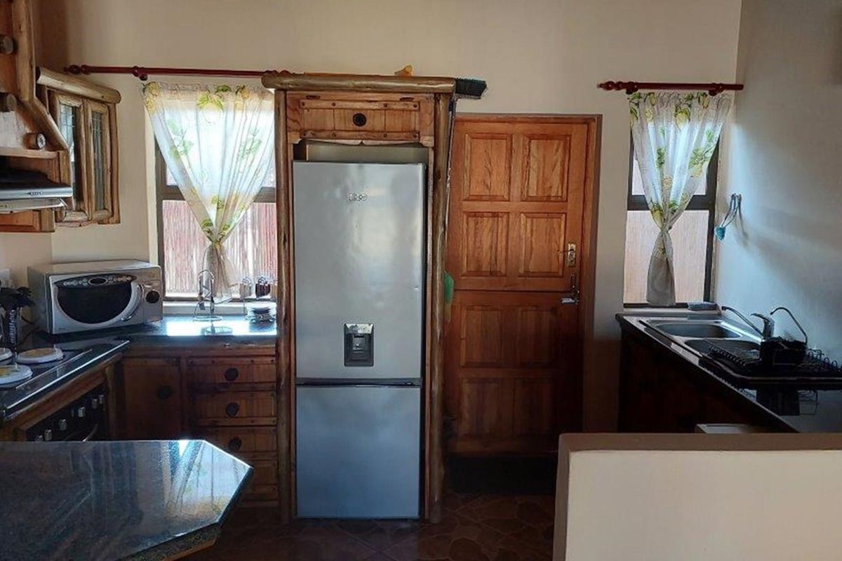 A functional kitchen featuring rustic wooden cabinetry, granite countertops, double sink, and integrated storage space for a refrigerator.