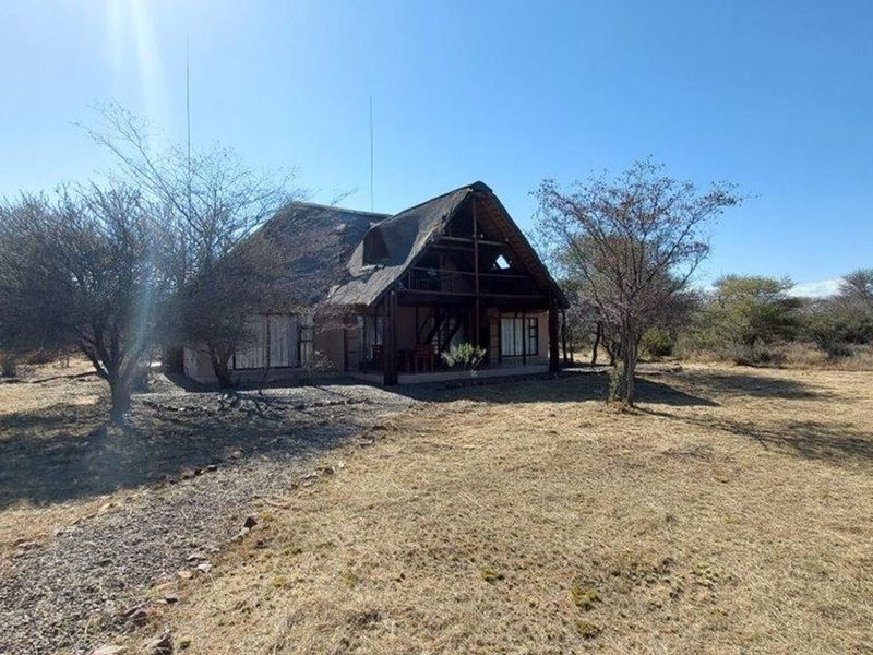 A rustic A-frame house with a thatched roof, featuring a covered patio and balcony, set within a natural, dry landscape.