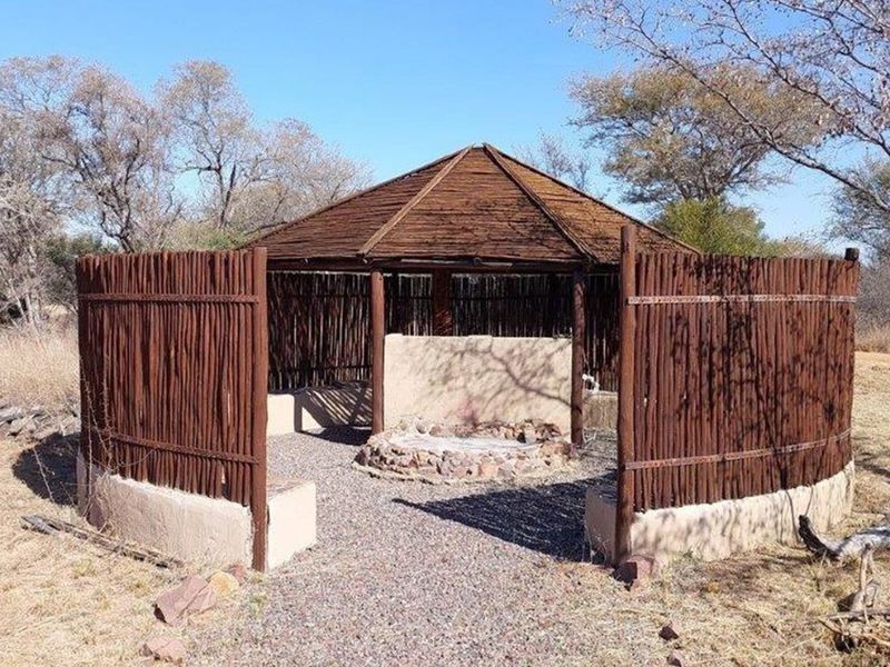An outdoor boma or entertainment area featuring a thatched roof structure, timber pole fencing, and a central stone-built fire pit in a natural setting.