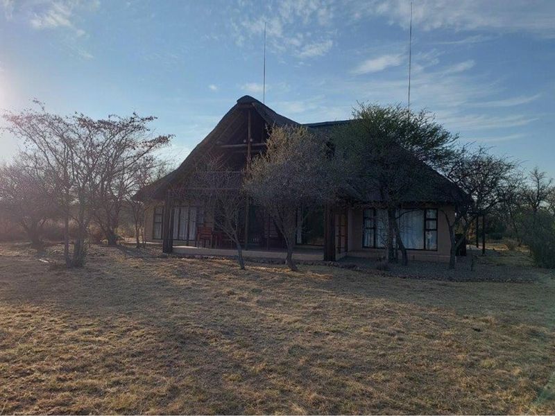 A rustic thatched-roof house exterior set within a natural bushveld landscape, featuring a covered patio and large windows.