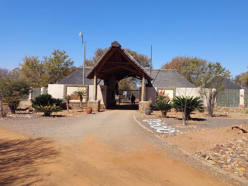 A rustic gated entrance featuring a traditional thatched roof structure, gravel driveway, and landscaped garden with indigenous plants.