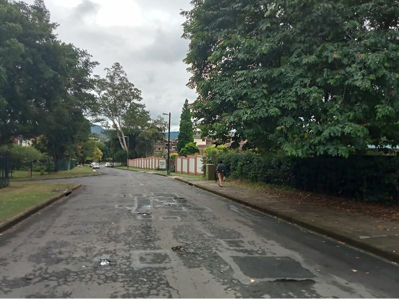 A view of the residential street approach featuring mature trees, perimeter walling, and a paved road surface with visible wear.
