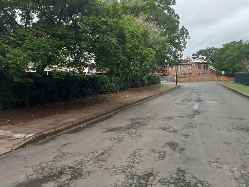 A street view showing the approach to the property with a paved road, sidewalk, and established boundary wall and trees.
