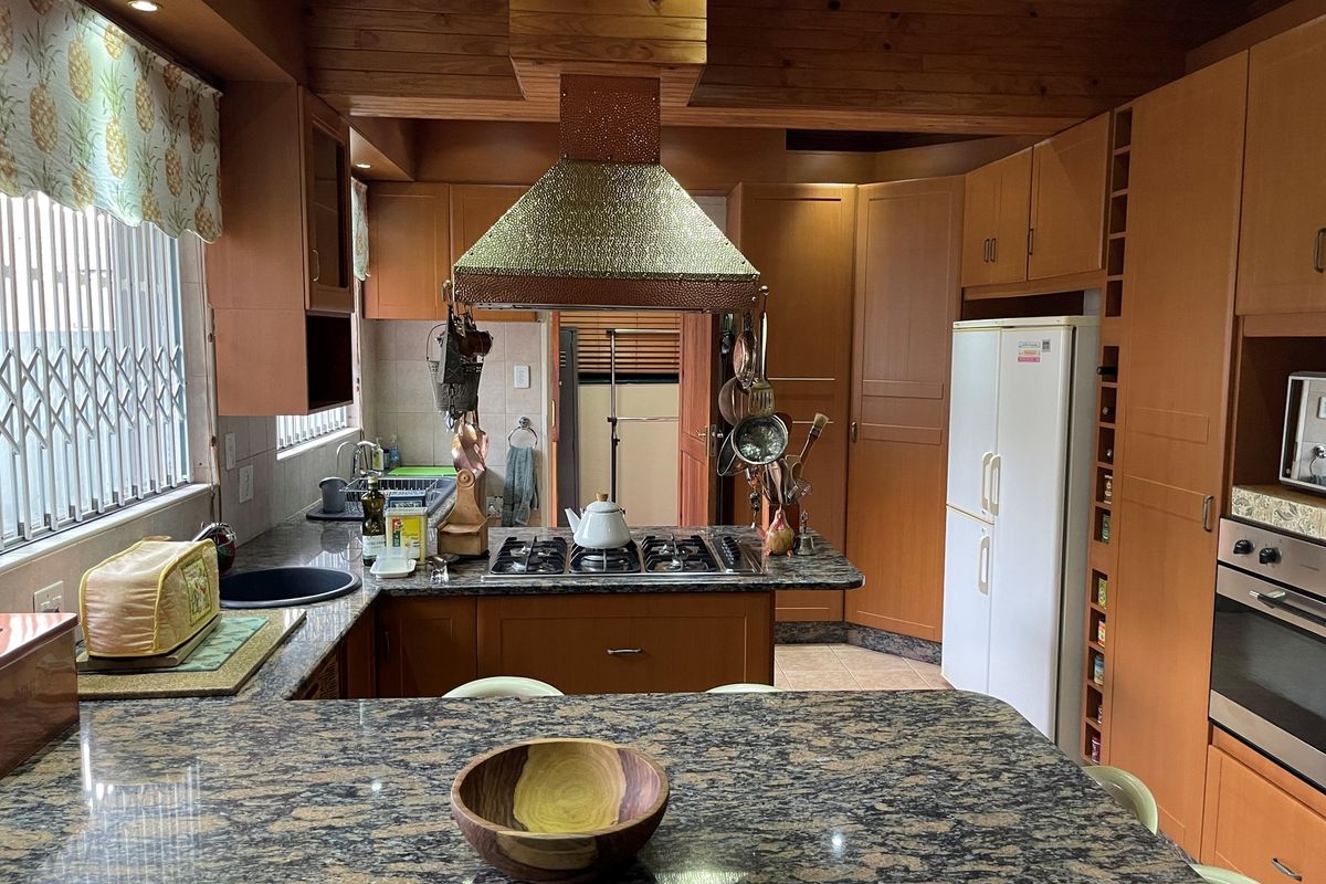 A traditional kitchen featuring warm-toned wooden cabinetry, granite countertops, a central island with a gas hob, and a distinctive copper-finish extractor fan.