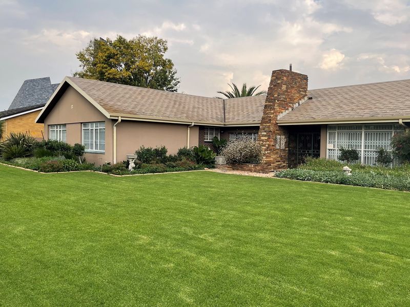 A well-maintained single-story house featuring a stone-clad chimney, shingle roof, and expansive front lawn with established garden beds.