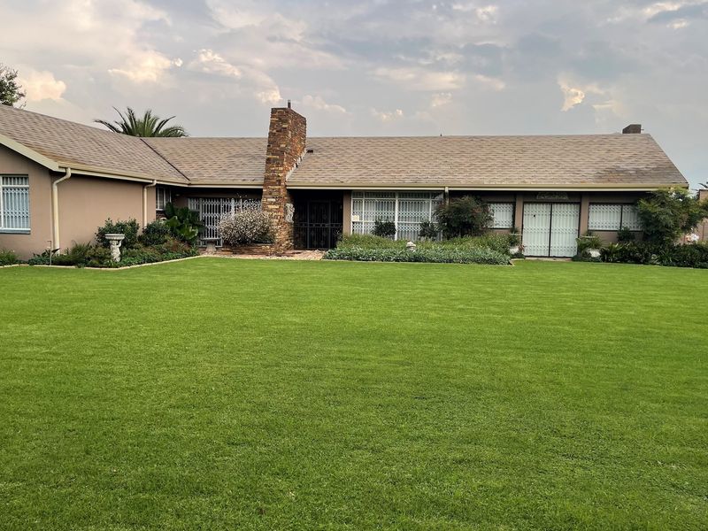 A well-maintained single-story residential home featuring a large manicured lawn, stone chimney, and secure window grilles.