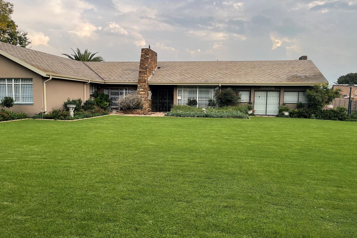 A well-maintained single-story residential home featuring a large manicured lawn, stone chimney, and secure window grilles.