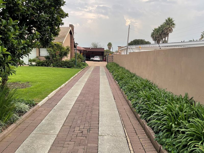 A long paved driveway leading to a rear carport, flanked by a well-maintained lawn and landscaped garden borders.