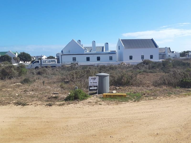 A vacant residential plot of land featuring natural coastal vegetation, with established white-walled residential properties visible in the background.