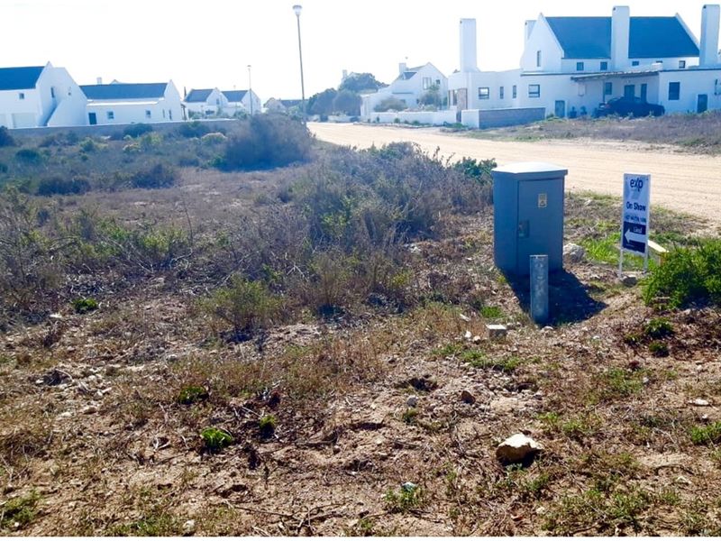 Vacant residential land plot featuring natural vegetation, an electrical utility box, and street access in a developed coastal estate.
