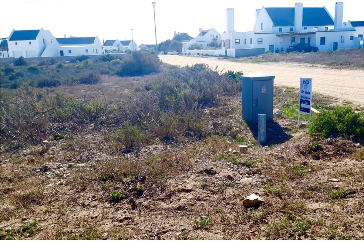 Vacant residential land plot featuring natural vegetation, an electrical utility box, and street access in a developed coastal estate.