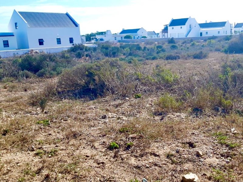 A vacant residential plot featuring natural vegetation, situated in a developed neighborhood with established white-walled homes in the background.
