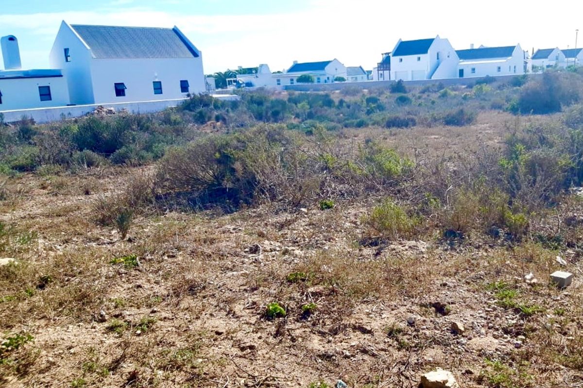A vacant residential plot featuring natural vegetation, situated in a developed neighborhood with established white-walled homes in the background.