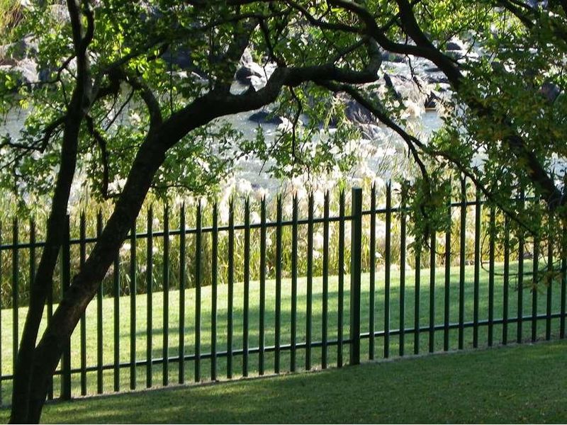 A view of the property's landscaped garden featuring a secure metal perimeter fence and mature trees overlooking a natural water feature.