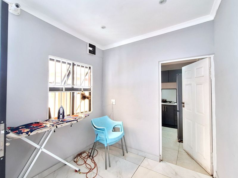 A clean utility space featuring tiled flooring, recessed lighting, and a window with security bars, providing access to the adjacent kitchen area.