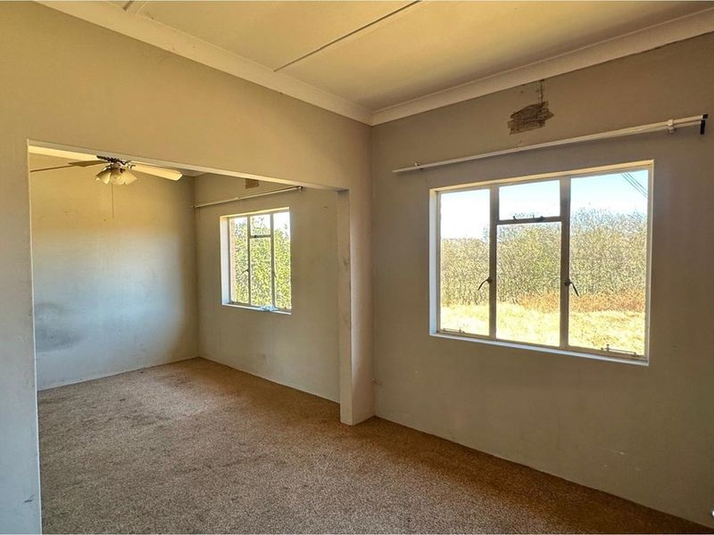 An empty, open-plan living area featuring carpeted flooring, a ceiling fan, and large windows, requiring cosmetic updates and repairs to the walls and ceiling.
