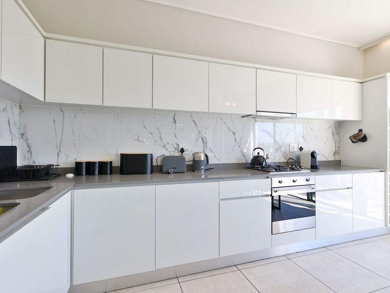 Modern kitchen featuring white gloss cabinetry, marble-look splashback, stone countertops, and integrated stainless steel appliances with tiled flooring.