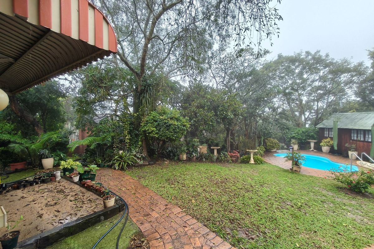 A spacious rear garden featuring a swimming pool, brick pathways, and a covered patio area, surrounded by mature trees and established landscaping.