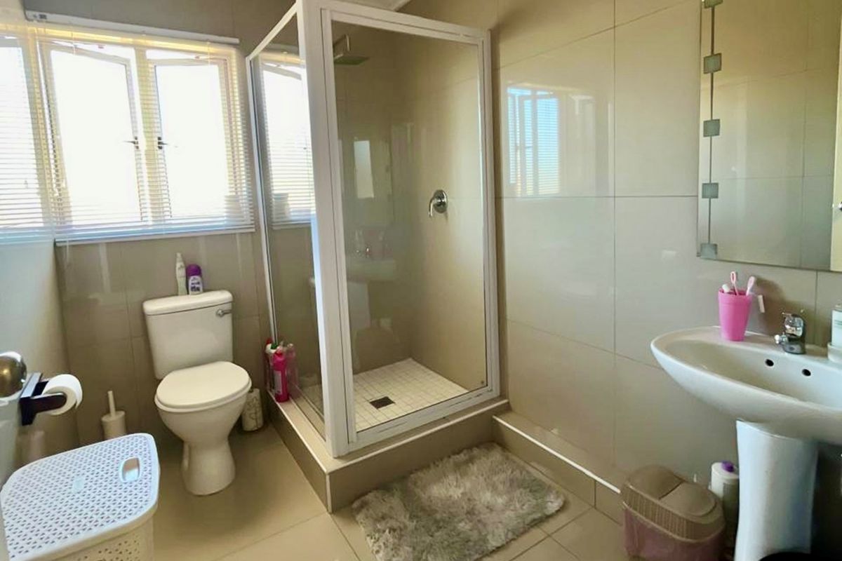 A functional bathroom featuring a glass-enclosed shower, pedestal basin, and toilet with neutral wall tiling and natural light from the window.
