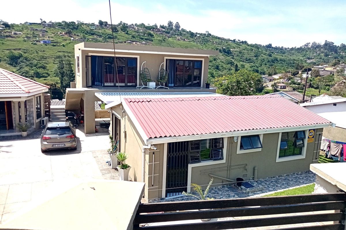 An elevated view of the property featuring a multi-level house with a red corrugated metal roof, paved driveway, and surrounding hillside landscape.