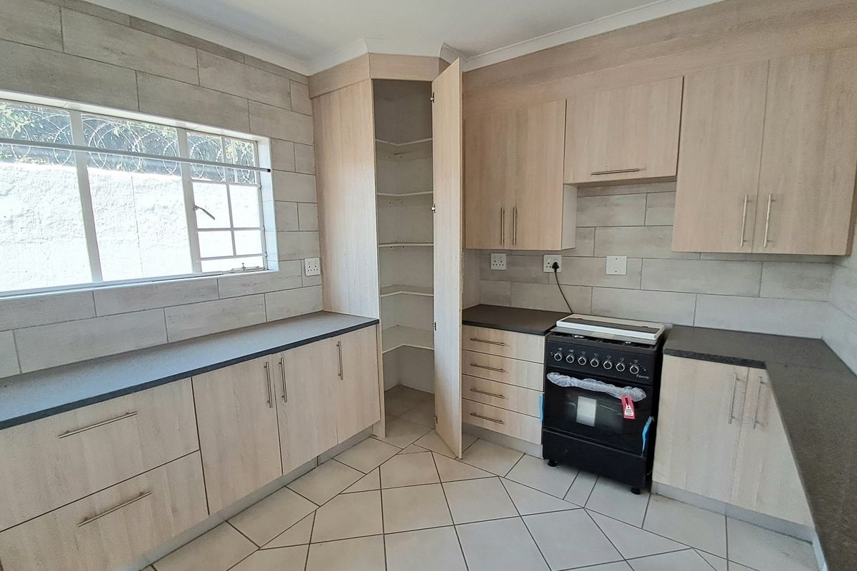 A clean, modern kitchen featuring light wood-finish cabinetry, dark countertops, a corner pantry, and tiled flooring.