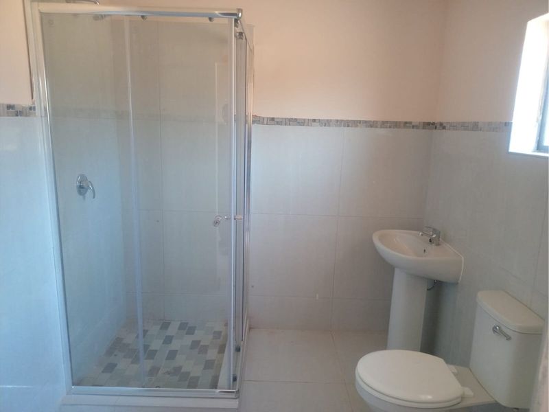A functional bathroom featuring a glass-enclosed shower, pedestal basin, and toilet with neutral wall tiling and a decorative mosaic border.