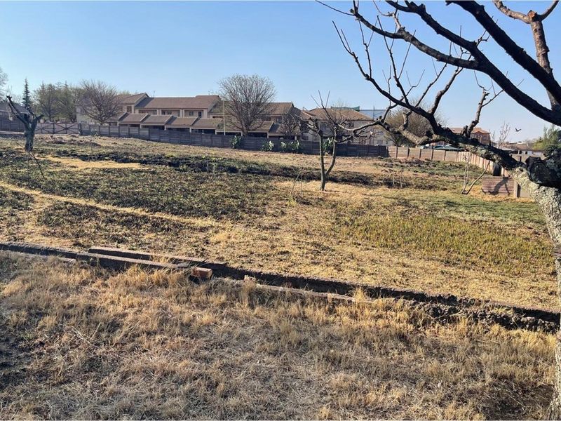 A view of a vacant residential plot featuring dry grass, scattered trees, and perimeter fencing, situated in a developed neighborhood.