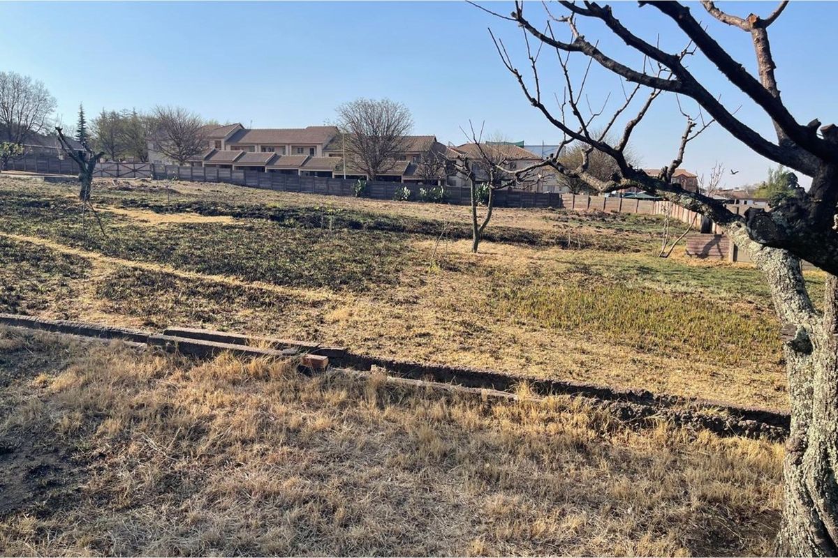 A view of a vacant residential plot featuring dry grass, scattered trees, and perimeter fencing, situated in a developed neighborhood.