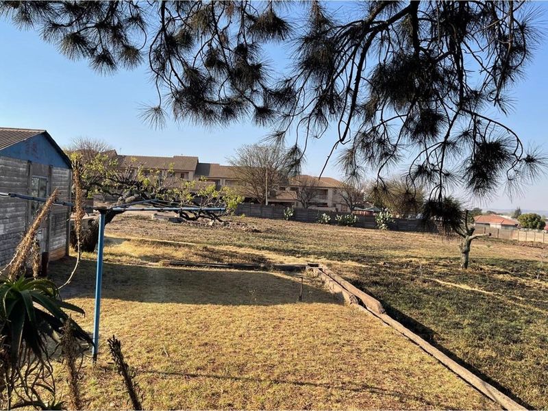 A view of a vacant residential plot featuring dry grass, mature trees, and an existing shed structure on the property.