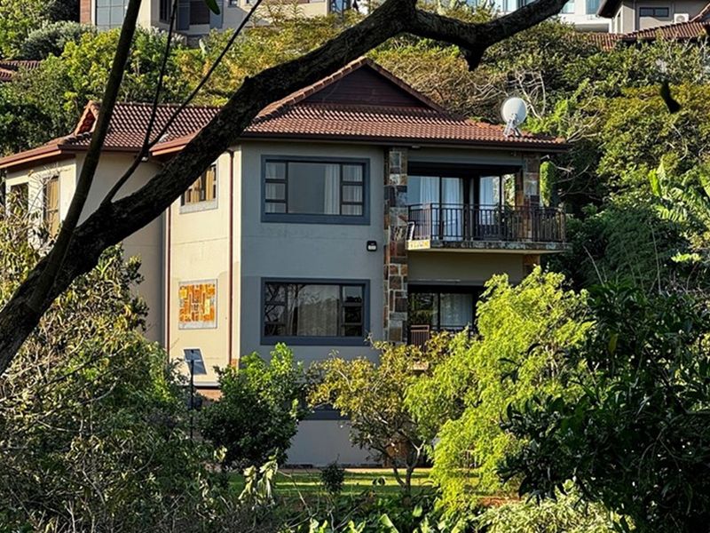 A multi-story residential property featuring a tiled roof, stone-clad accents, and balconies, nestled within a lush, mature garden setting.