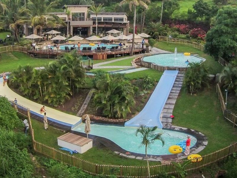 Aerial view of a recreational pool area featuring multiple swimming pools, a water slide, extensive landscaped gardens, and a large wooden deck.