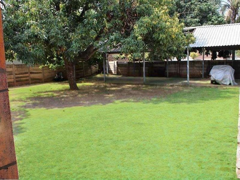 Spacious rear garden featuring a large lawn, mature trees, and a covered carport structure with a corrugated metal roof.