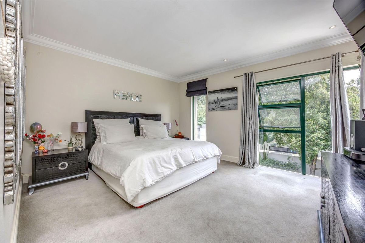 A spacious bedroom featuring neutral carpeting, crown molding, and large windows with green frames that provide natural light and views of the surrounding greenery.