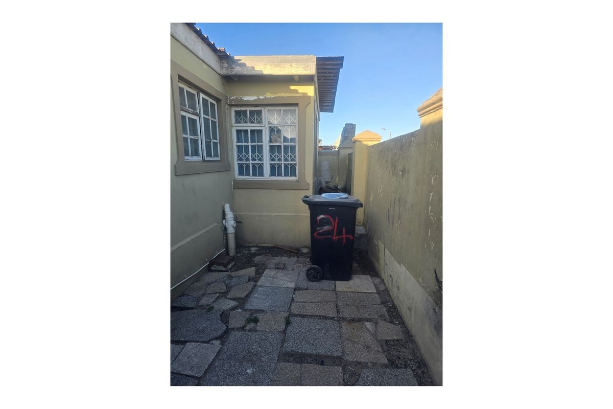 A narrow rear exterior area featuring stone paving, a boundary wall, and windows with security bars, showing signs of wear and maintenance needs.