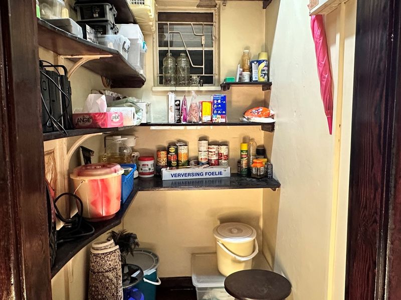 A compact walk-in pantry or scullery area featuring built-in wall-mounted shelving and a small window with security bars.