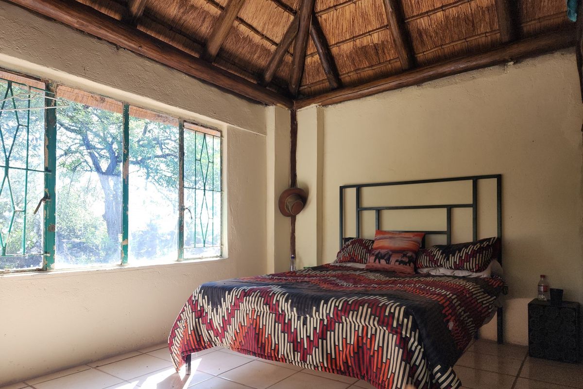 A rustic bedroom featuring a traditional thatched roof with exposed timber beams, tiled flooring, and large windows overlooking the surrounding farm landscape.