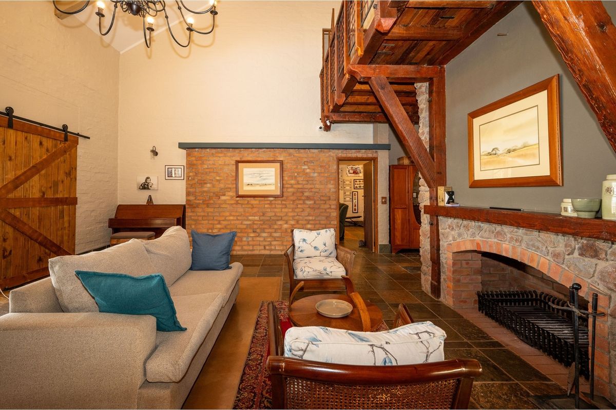 Spacious living room featuring a double-volume ceiling, exposed timber mezzanine, rustic stone fireplace, and brick feature wall with slate flooring.
