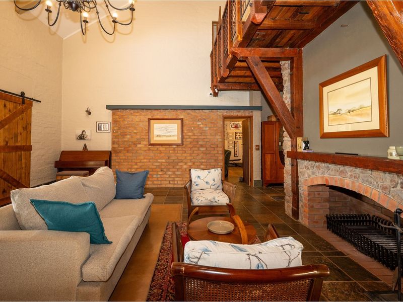 Spacious living room featuring a double-volume ceiling, exposed timber mezzanine, rustic stone fireplace, and brick feature wall with slate flooring.