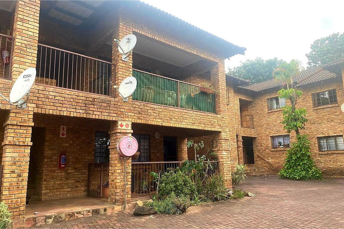 A multi-story apartment complex featuring a face brick facade, tiled roof, and paved courtyard with metal balcony railings.