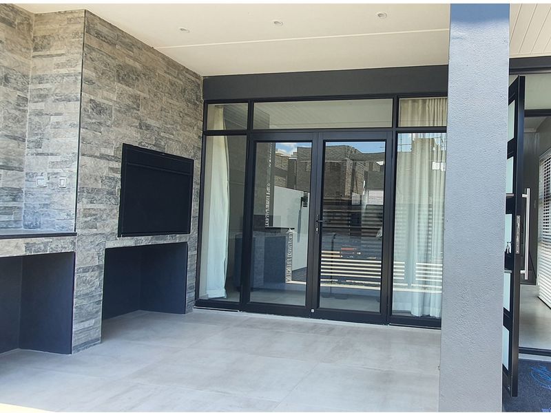 Covered patio featuring a built-in braai with stone cladding, tiled flooring, and large glass sliding doors providing seamless access to the interior living space.