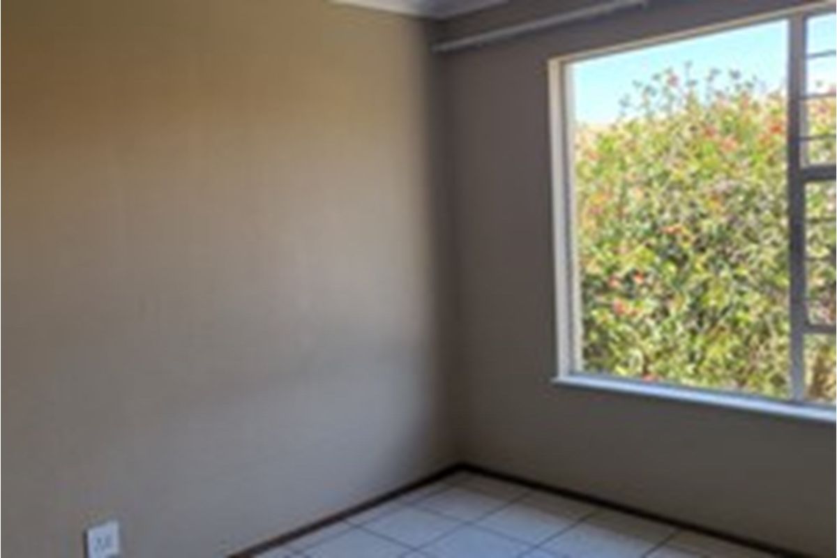 A bright, empty bedroom featuring tiled flooring, neutral wall paint, and a large window overlooking garden greenery.