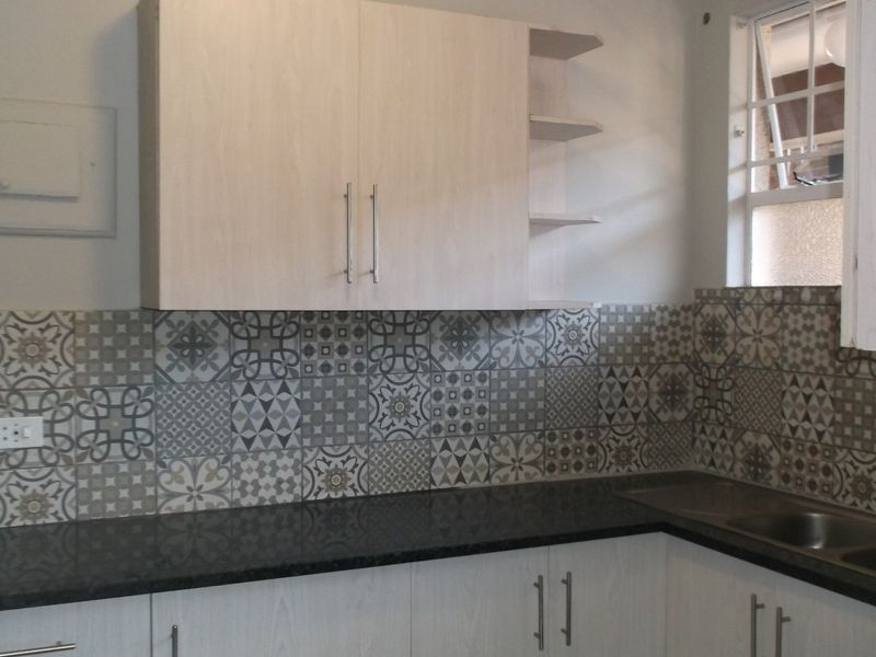 A clean, modern kitchen featuring light wood-tone cabinetry, dark granite countertops, and a decorative patterned tile backsplash with a functional stainless steel sink.