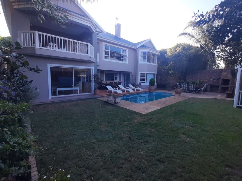 Rear view of the property featuring a swimming pool, paved patio area, built-in braai, and an upper-level balcony overlooking the garden.